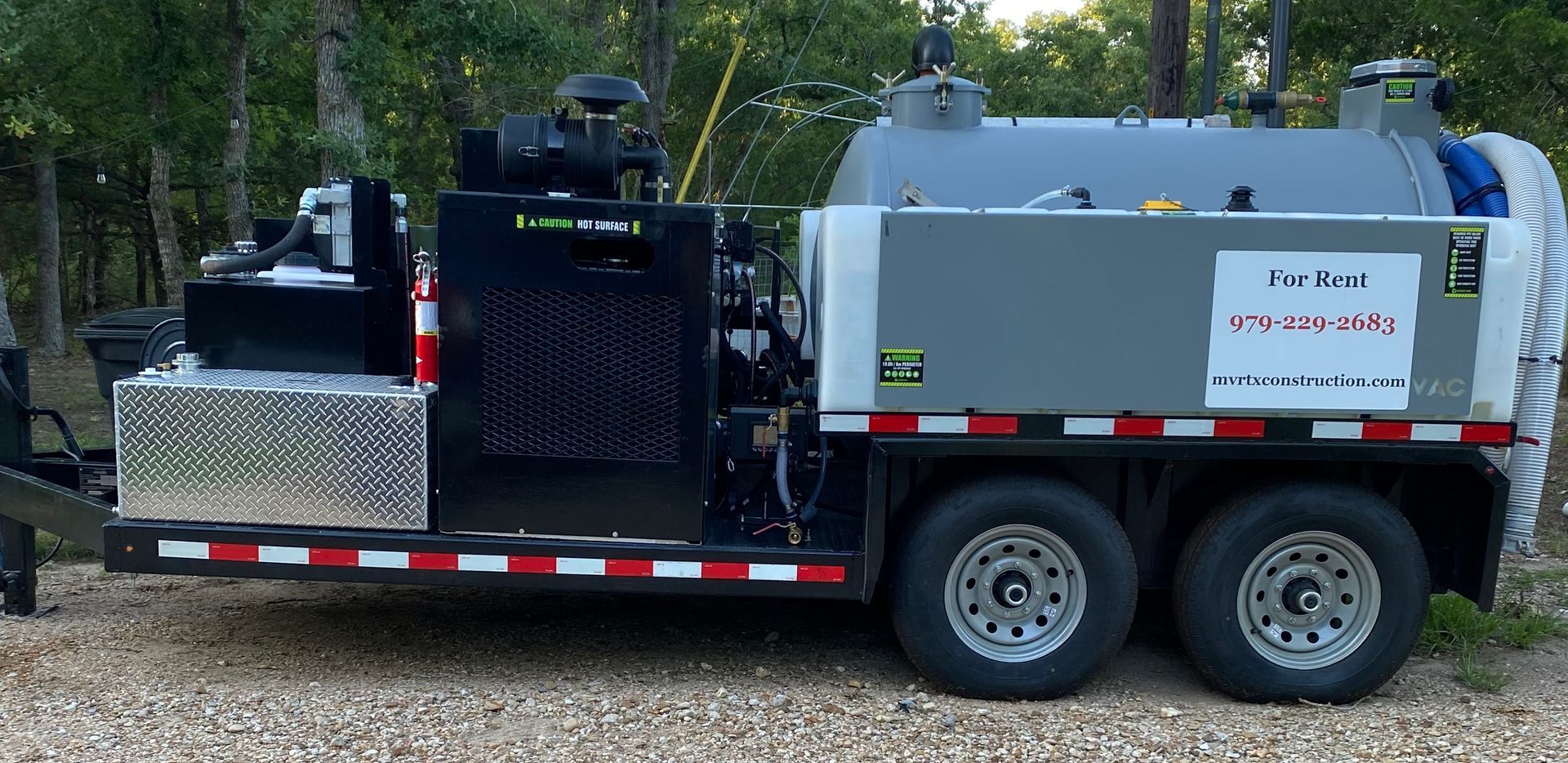 A trailer with a large tank on the back is parked on gravel.