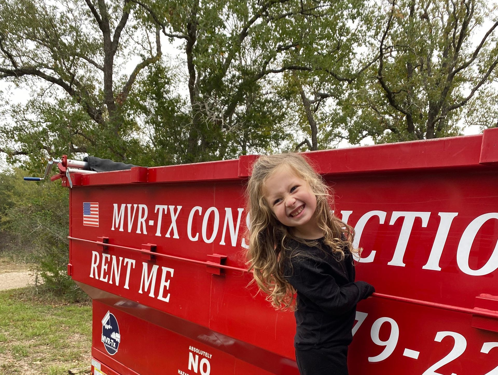 A little girl is standing in front of a red dumpster that says rent me
