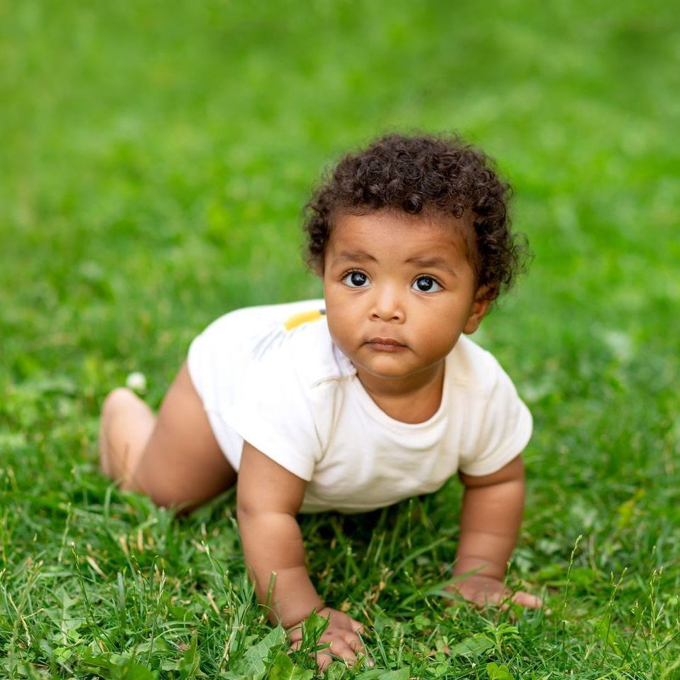 A small child is crawling on the grass and looking up at the camera. 