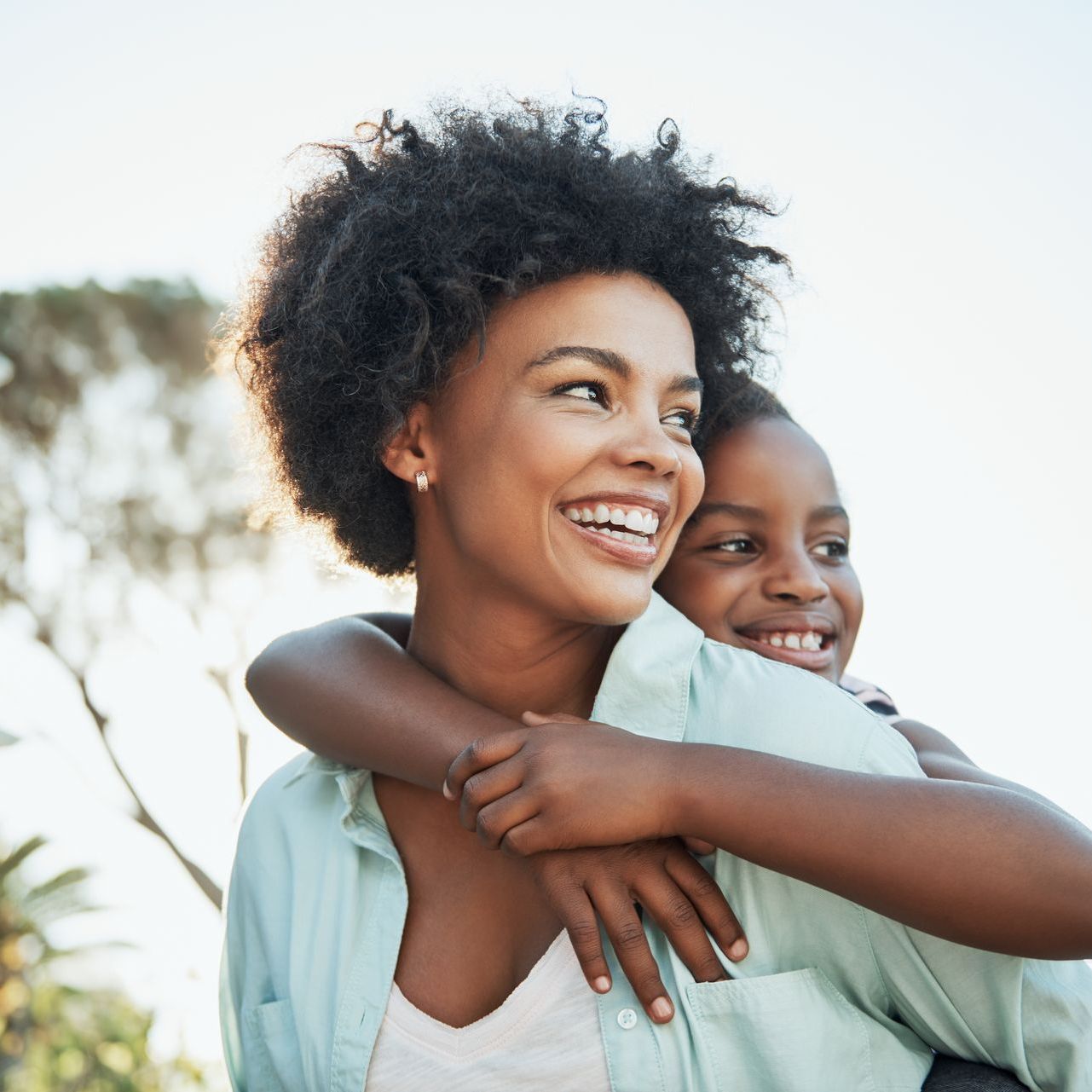 Smiling woman with child hugging her from behind. They are outside with a tree behind them.