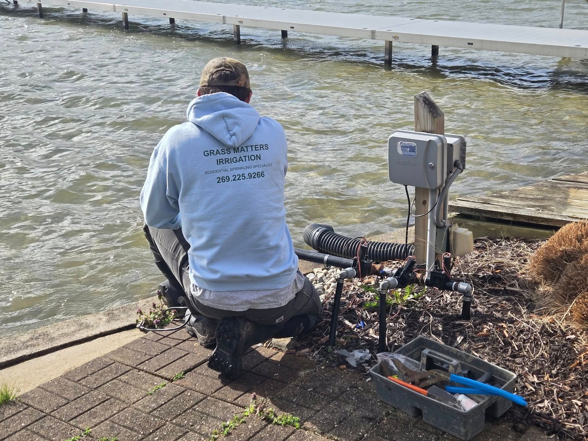 Man in a light blue sweatshirt kneels, working on equipment near water's edge, next to a control box.