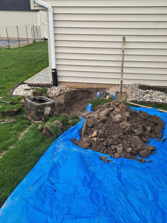 Hole dug near house foundation. Dirt on blue tarp, shovel nearby. Downspout and small stone border visible.