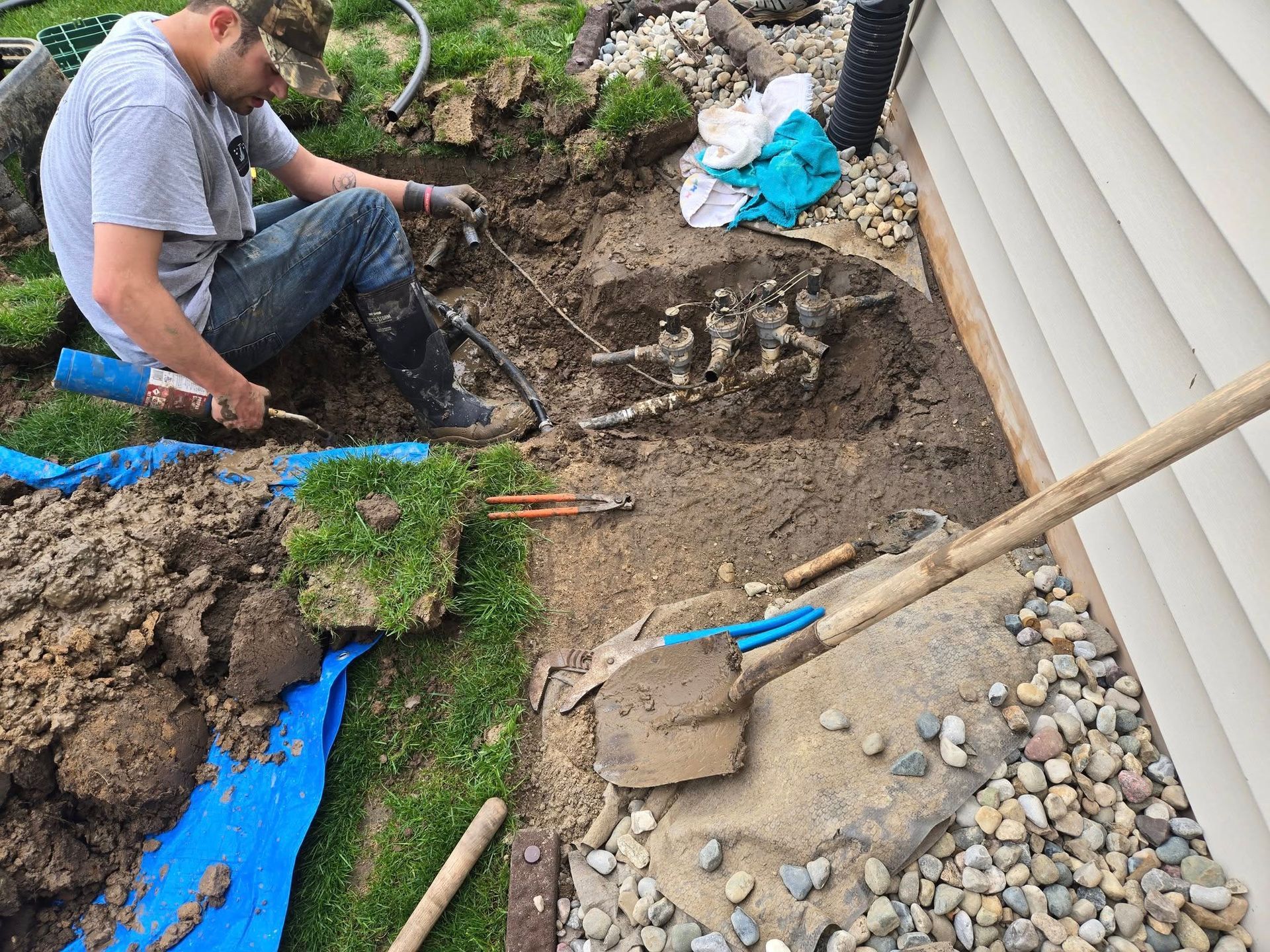 Man digging in yard near a house, exposed pipes, dirt, tools, and grass visible.