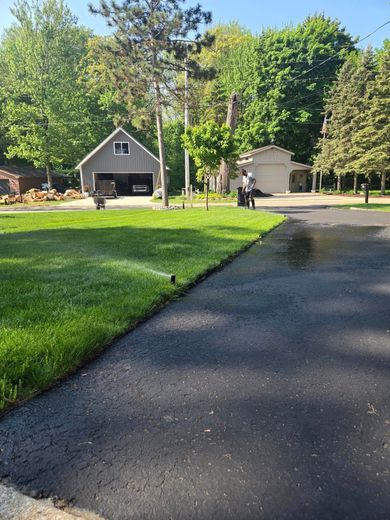 Lawn with sprinklers and asphalt driveway. A gray garage and trees are in the background under a blue sky.