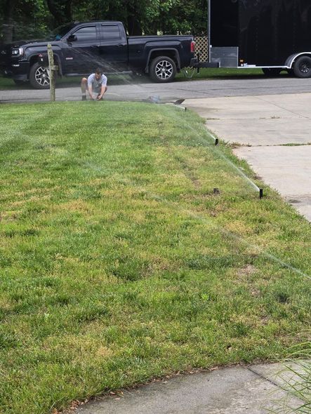Person crouches near lawn sprinklers spraying water onto a patchy green lawn with a truck and trailer in the background.