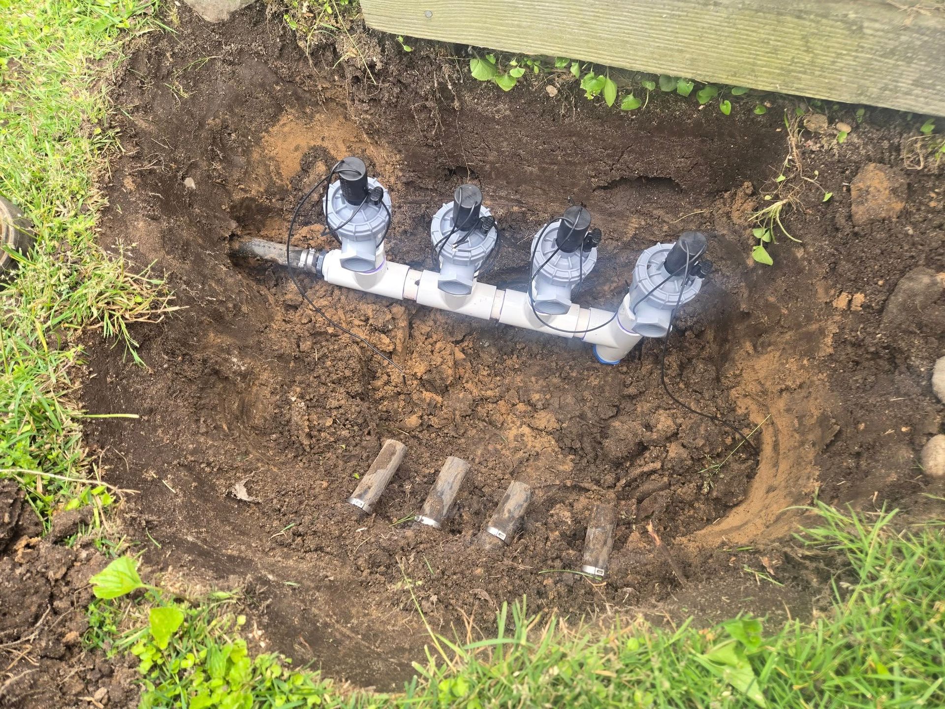 Irrigation system with four valves and exposed PVC pipes, installed in a garden bed.
