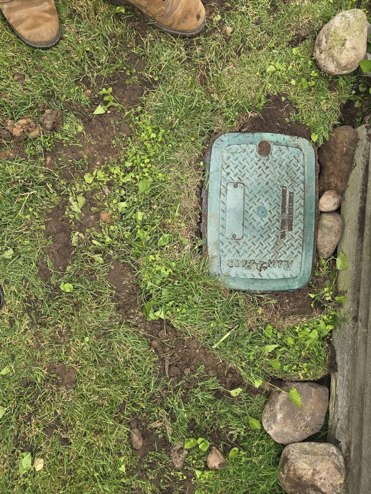 Green irrigation valve box in grass next to a stone border; dirt path visible.
