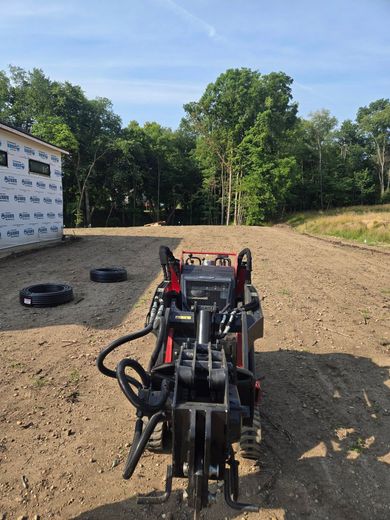 A red mini skid steer on a gravel surface, near a building under construction and trees on a sunny day.
