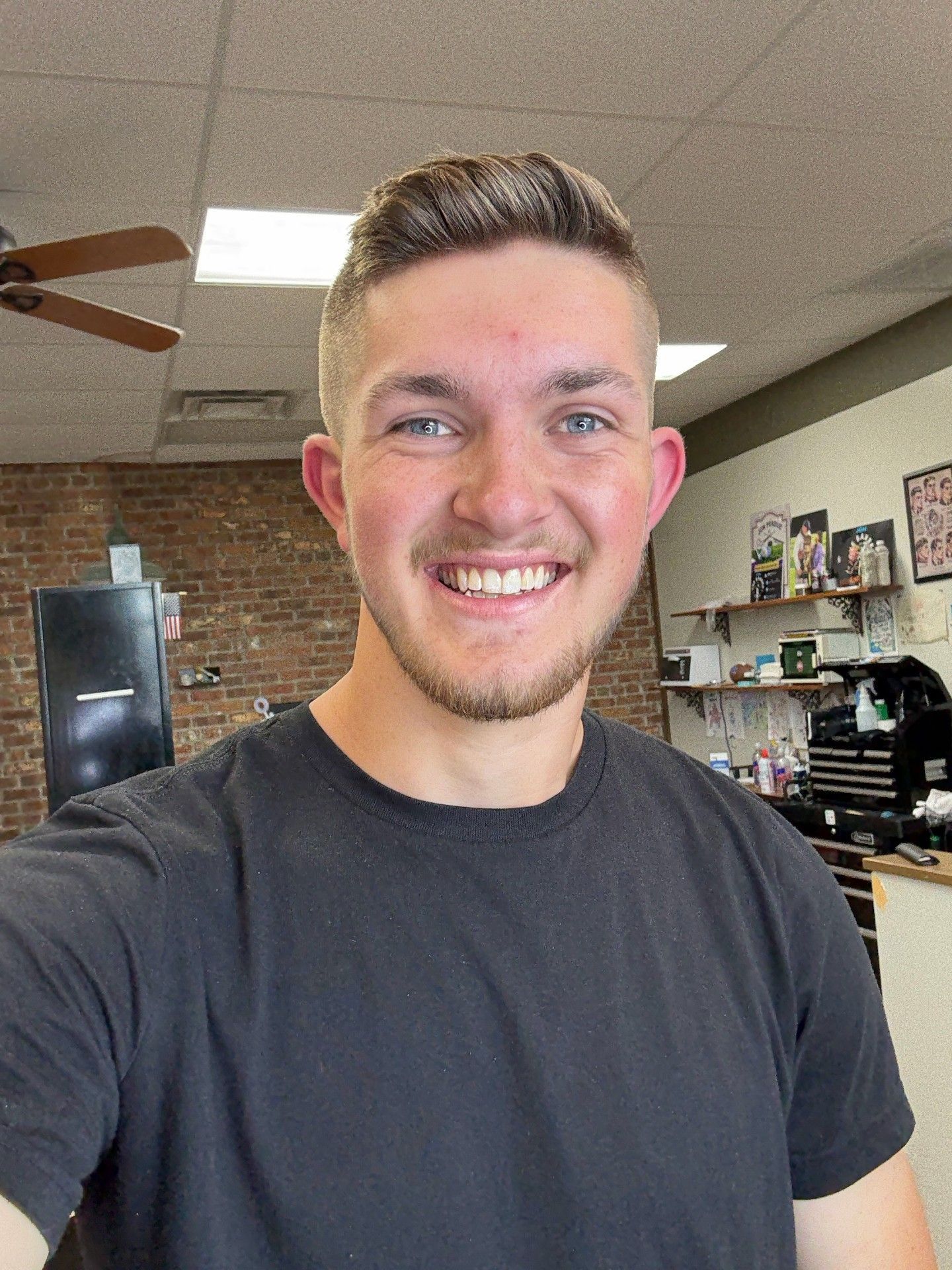 Man with a fade haircut smiles in a barbershop. He wears a black shirt, brick wall and ceiling fan visible behind him.