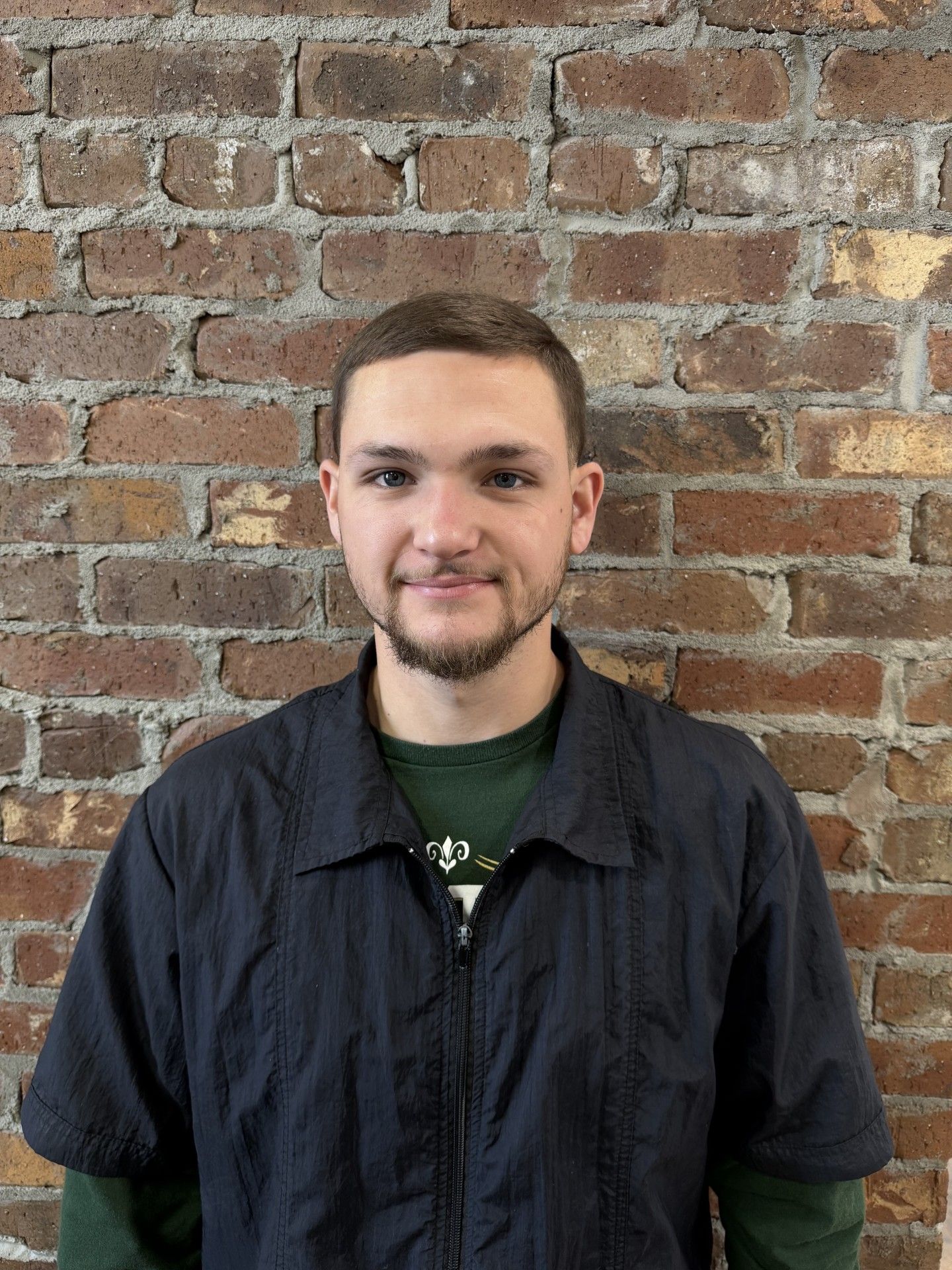 Man with short hair and stubble, wearing a dark jacket over a green shirt, standing in front of a brick wall.
