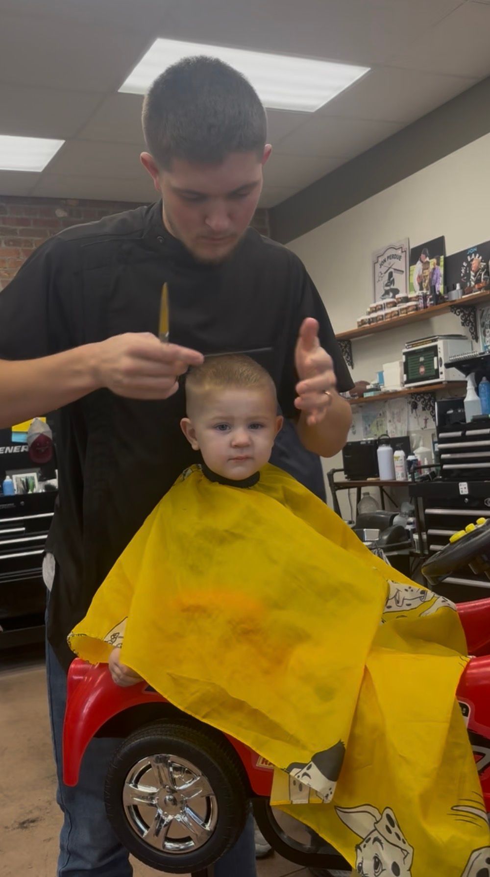 Barber giving a toddler a haircut in a shop. Child is in a red car chair, wearing a yellow cape.