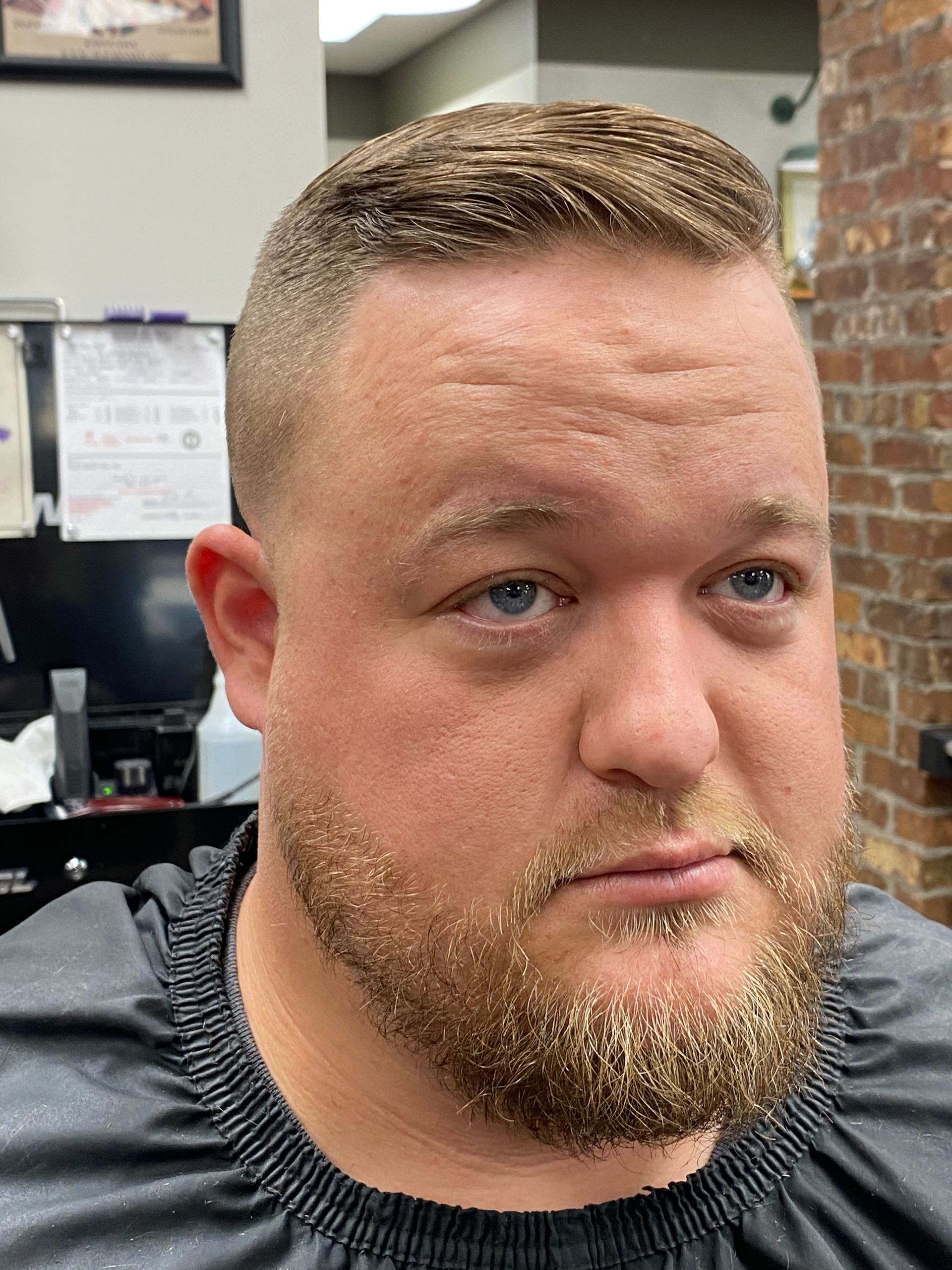 Man with light brown hair and beard, wearing a black barber's cape, in a barbershop.