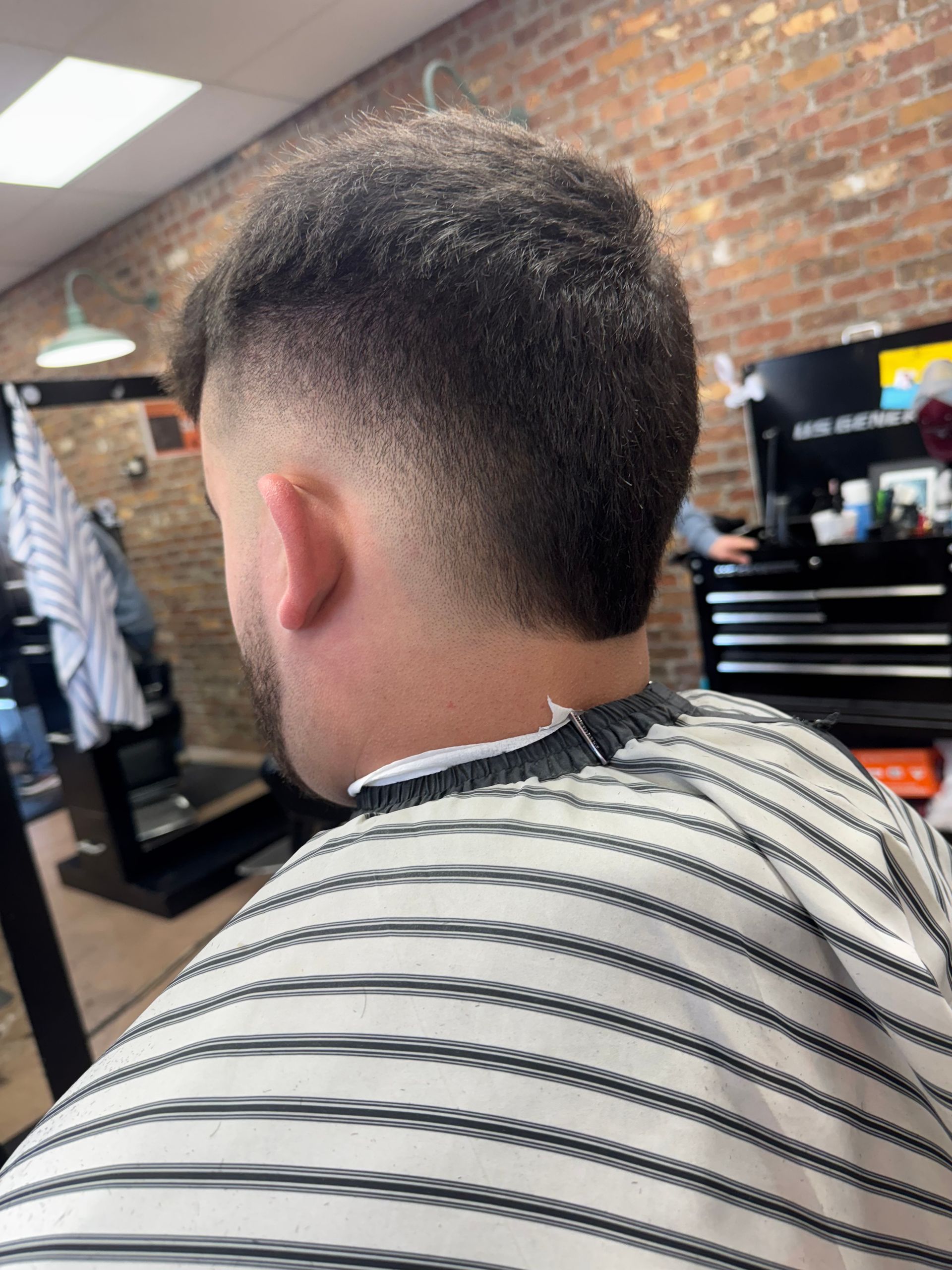 Man getting a haircut, blurred background of a barbershop. Faded short hair with striped barber cape.