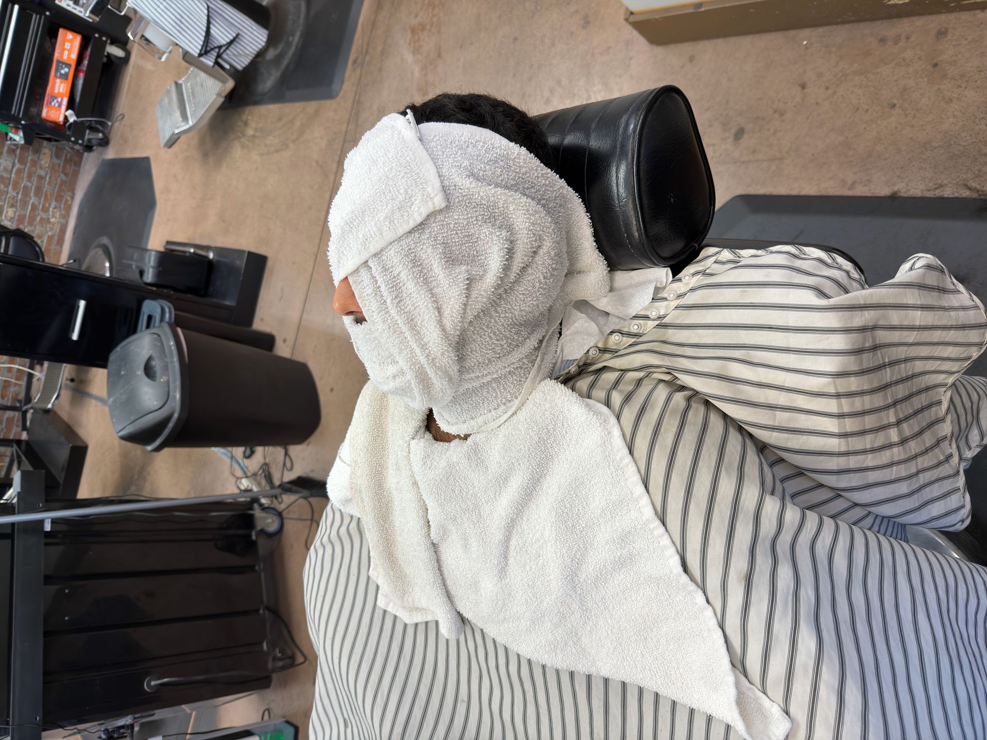Person at a barber shop, wrapped in towels. Sitting in a chair with a striped apron.