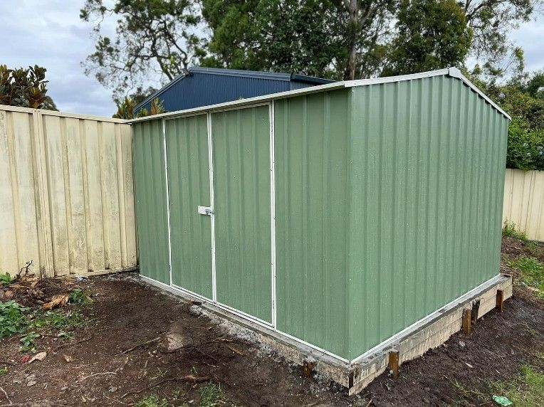 A gReen Metal Shed is Sitting in the Dirt Next to a Fence — JOA - Construction & Maintenance in Appin, NSW