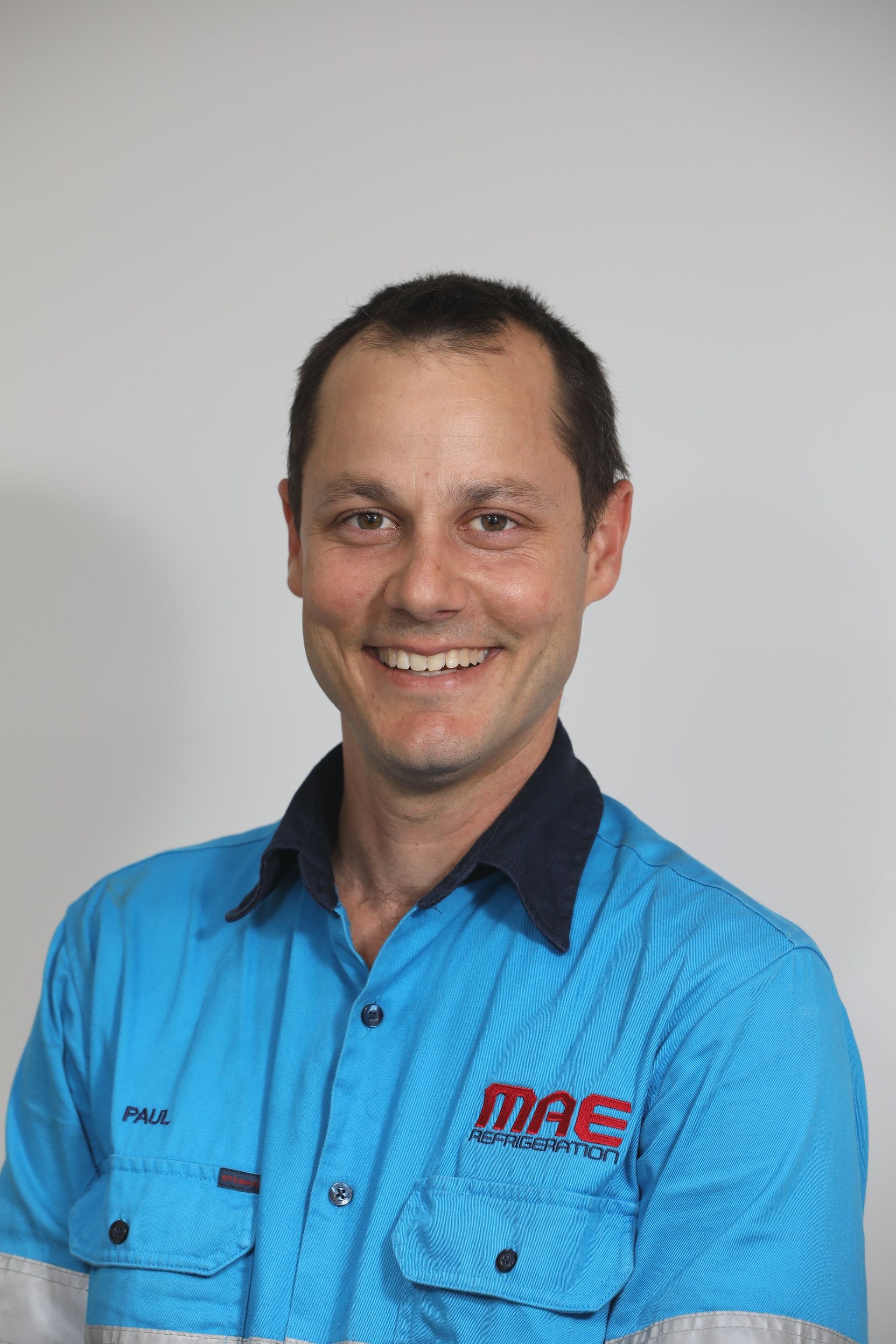 Man wearing a blue work shirt, smiling, against a plain background — MAE Refrigeration In Portsmith, QLD
