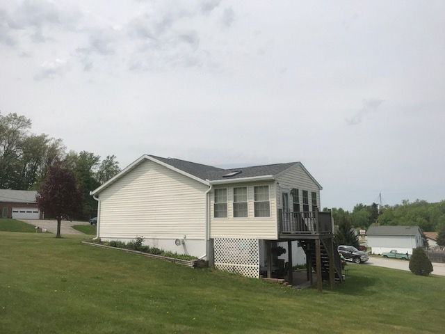 A light-colored house with a dark roof behind a chain-link fence and green bushes with pink roses.