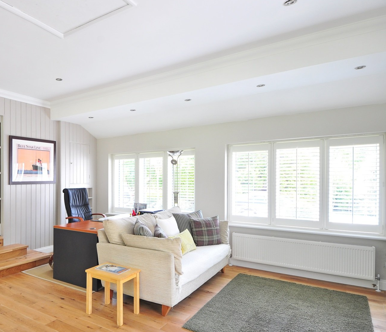 A living room featuring a light sofa with patterned cushions, a wooden side table, and a desk, set against white walls.