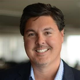 A headshot of a person with dark hair smiling at the camera, wearing a blue blazer over a light blue checkered shirt.