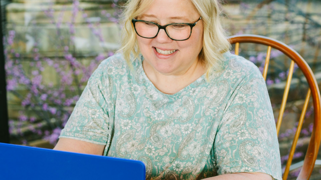 A person with blonde hair and glasses smiles while sitting in a wooden chair, typing on a bright blue laptop.