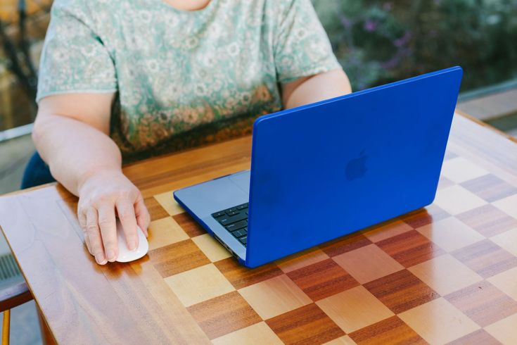 A person sits at a wooden checkerboard-patterned table using a white mouse next to a bright blue laptop.