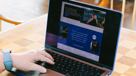 A person wearing a light blue watch types on a laptop, displaying a website with blue text blocks and imagery on a table.