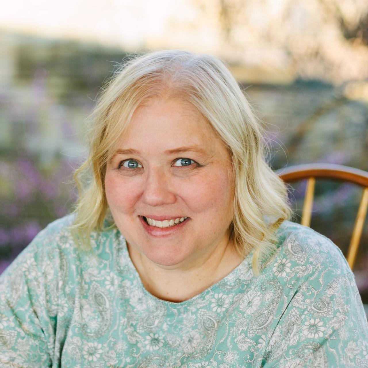 A smiling woman with blonde hair wearing a light blue floral top, positioned in front of an outdoor, blurred background.