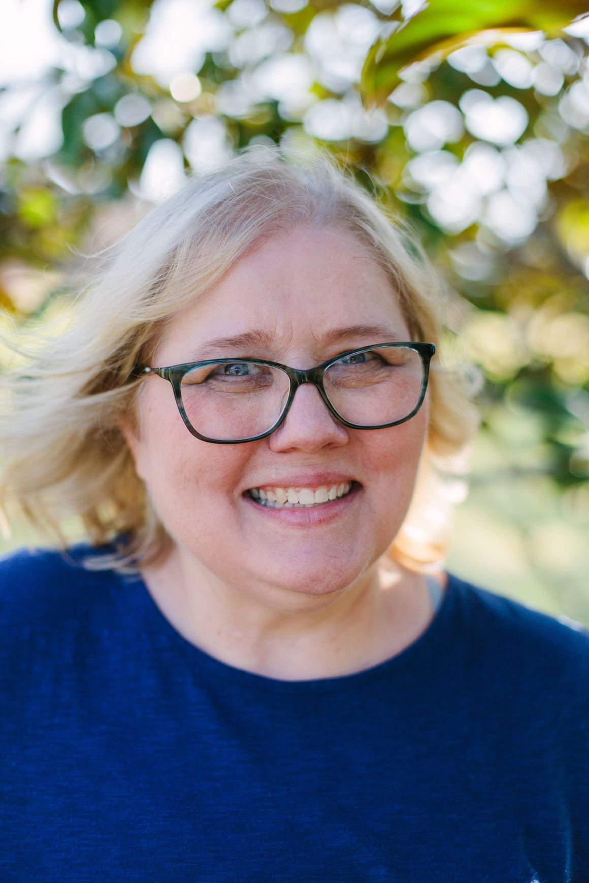 A smiling person with blonde hair and dark-rimmed glasses wearing a blue shirt, positioned outdoors with bokeh lighting.