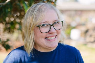 A person with shoulder-length blonde hair and glasses smiles broadly in front of a soft-focus outdoor background.