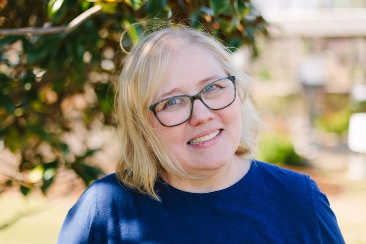 A smiling person with blonde hair and black-rimmed glasses wearing a navy blue top, posed outdoors near green foliage.