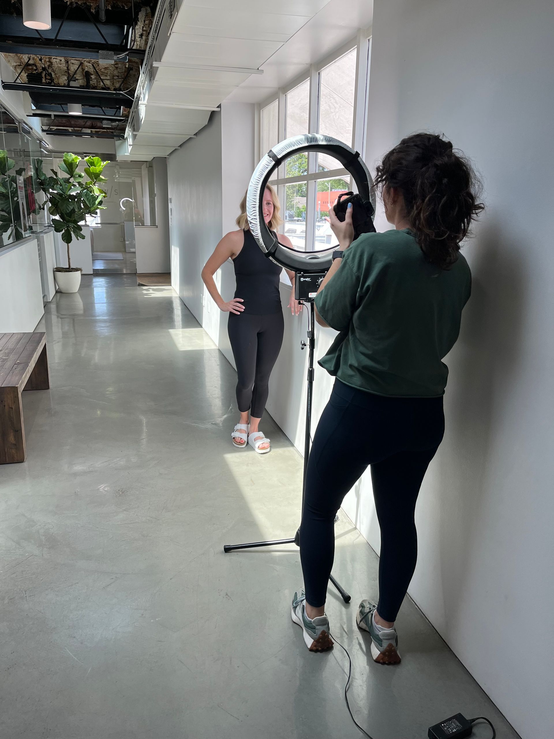 A person filming another individual standing in a bright, modern hallway using a ring light and camera.