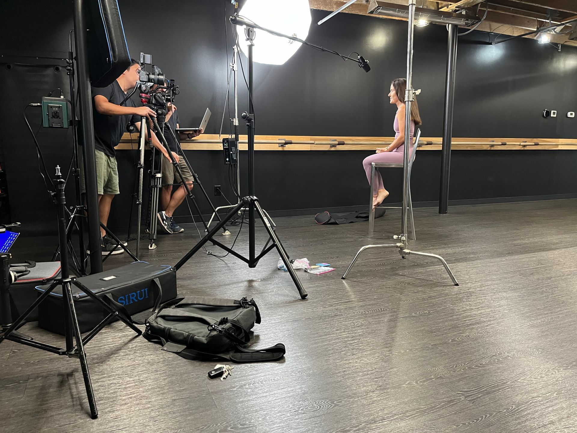 A videographer films a person sitting on a stool in a studio with professional lighting and black walls.