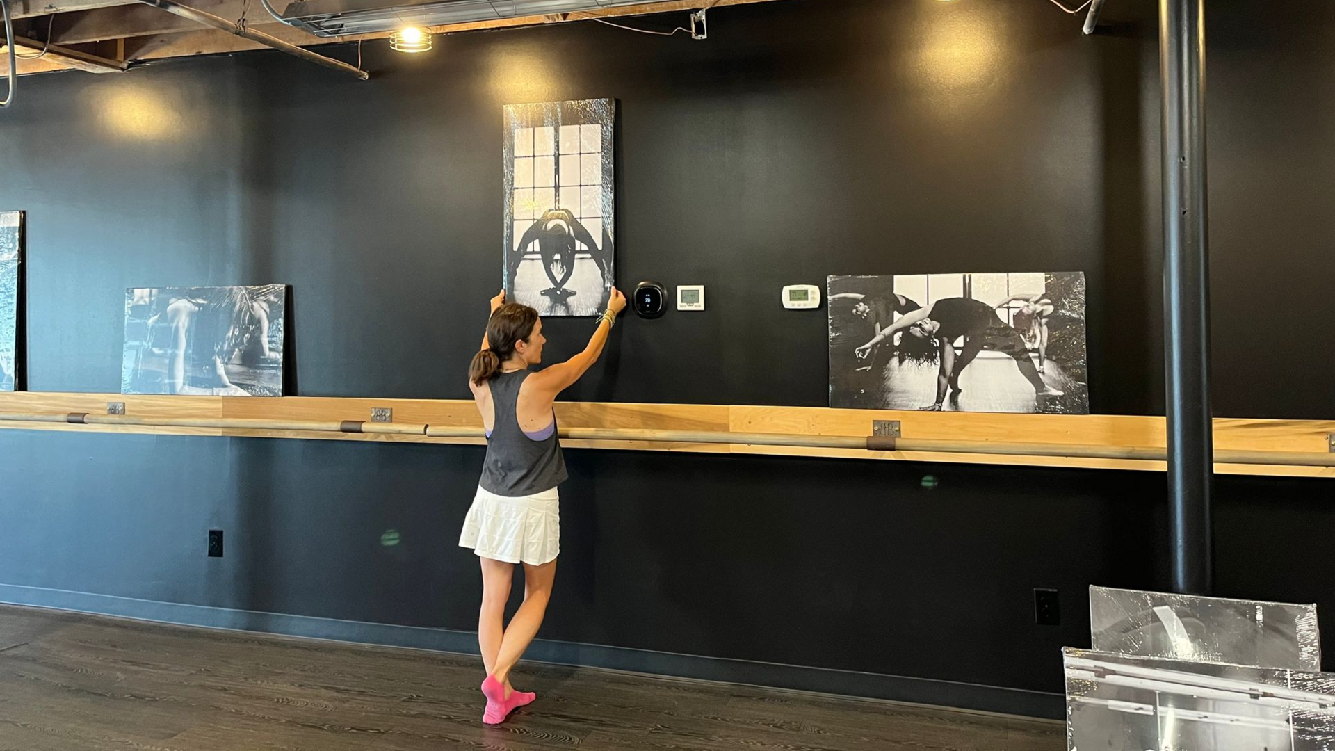 A person in a gray tank top and white skirt adjusts a vertical wall-mounted photograph in a room with black walls.