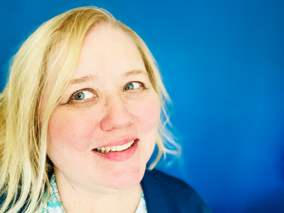 A person with blonde hair smiles softly in front of a solid blue background.