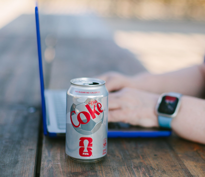 A can of Diet Coke sitting on a wooden table next to a person using a blue laptop and wearing a smartwatch.