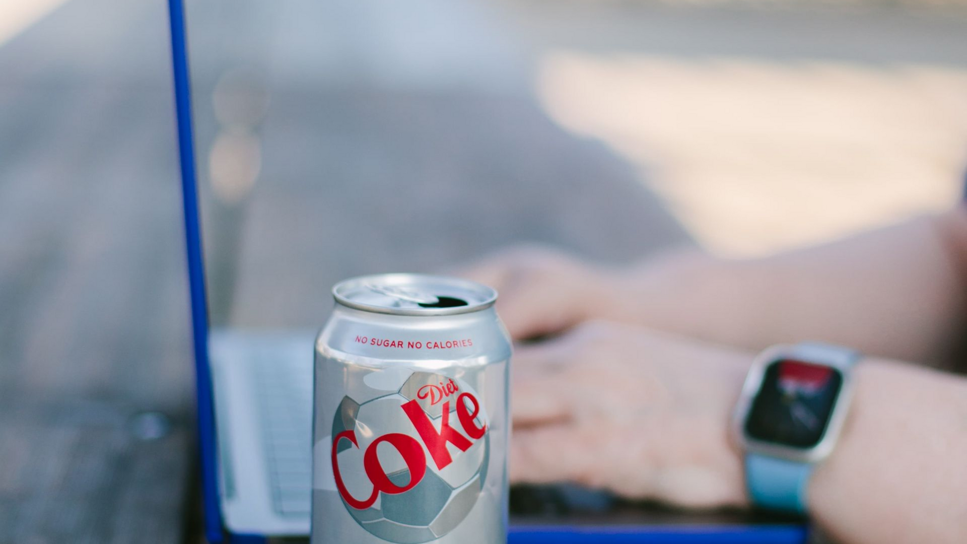 A Diet Coke can sits on a surface in front of a person working on a laptop while wearing a smartwatch.