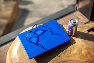 A bright blue laptop sits on a light-colored wooden table with a pair of eyeglasses on top and a soda can beside it.