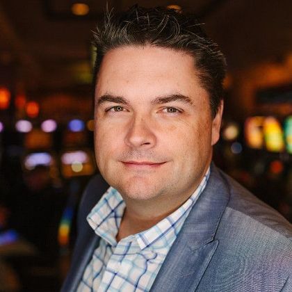 Headshot of a person in a blue blazer and light blue plaid shirt, set against a blurred background of casino slot machines.