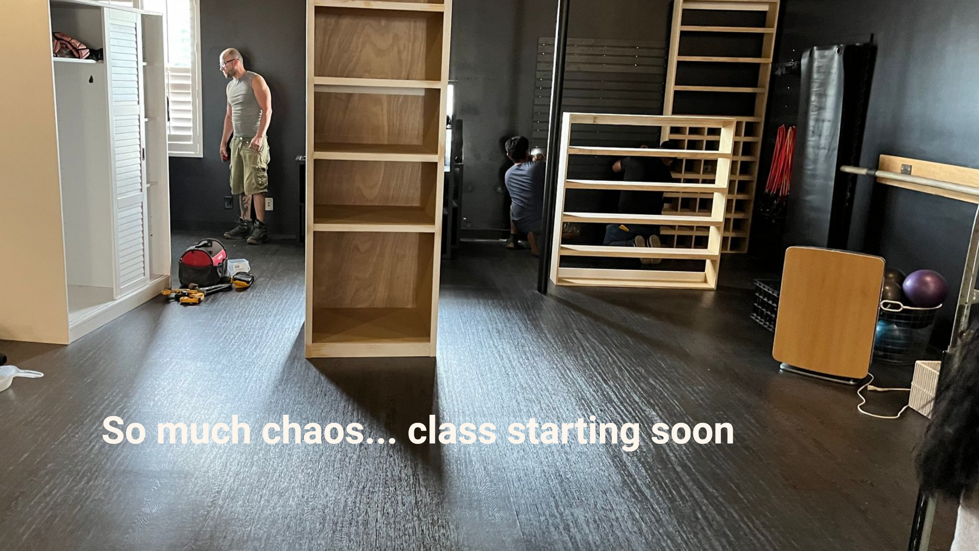 An interior room with dark flooring, wooden shelving units, and people preparing for a class.