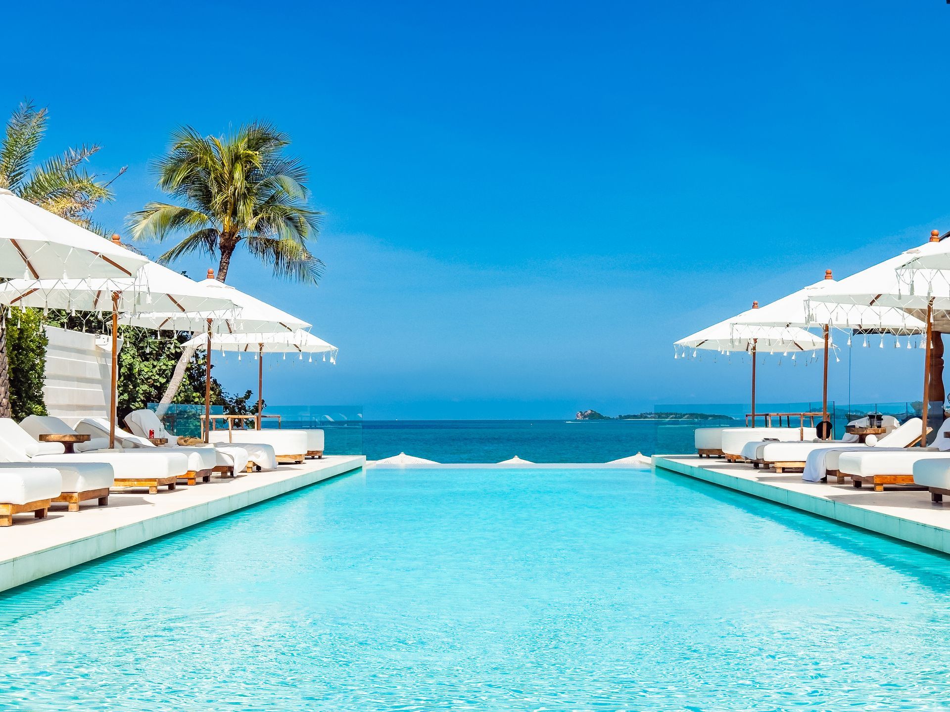 A bright, turquoise infinity pool surrounded by white lounge chairs and umbrellas, overlooking the blue ocean and sky.