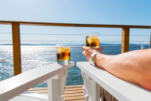 A person sits on white chairs on a deck, holding a glass of iced amber liquid while another glass sits on the chair arm.
