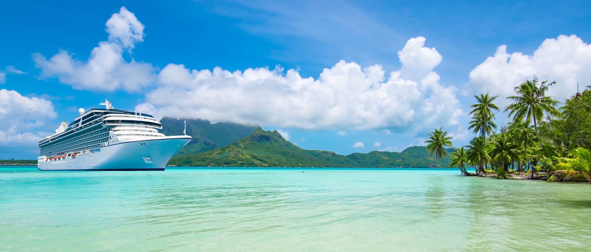 A large cruise ship floats on turquoise tropical waters near a lush green island with palm trees under a blue, cloudy sky.