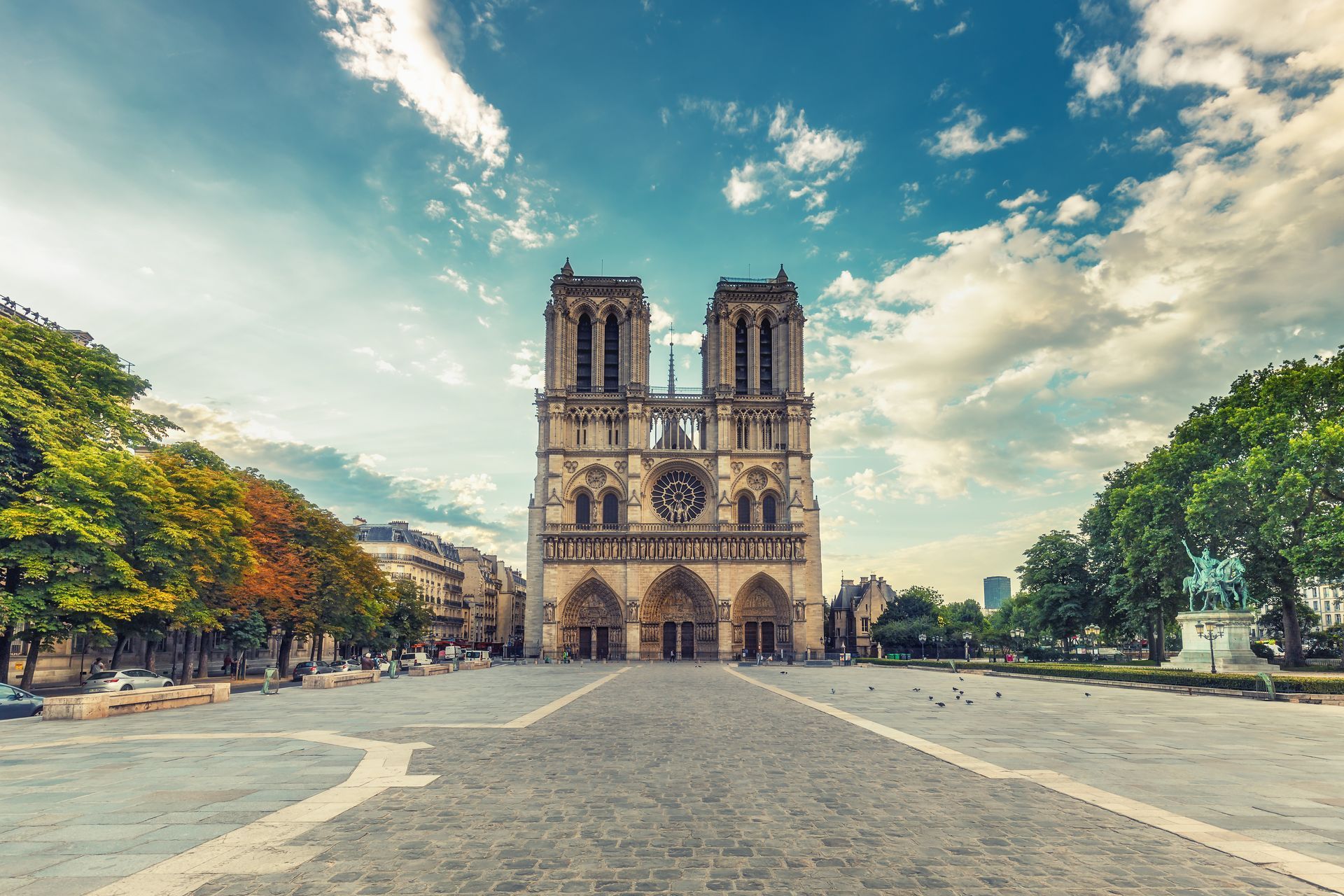 Front view of Notre-Dame Cathedral in Paris under a blue sky with scattered clouds, surrounded by trees and a stone plaza.