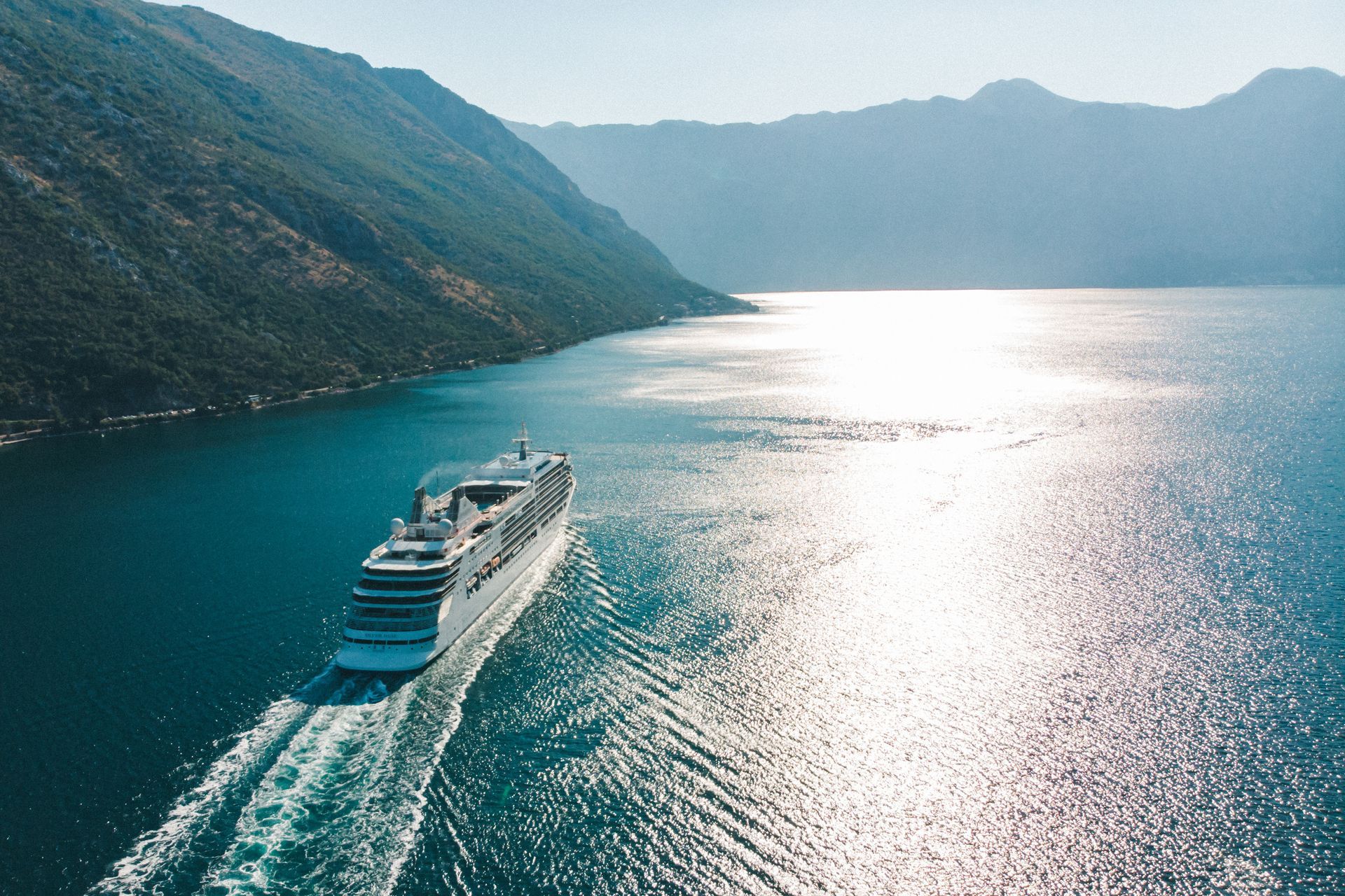 A large white cruise ship sails through a calm, sun-dappled bay surrounded by steep, grassy mountains.