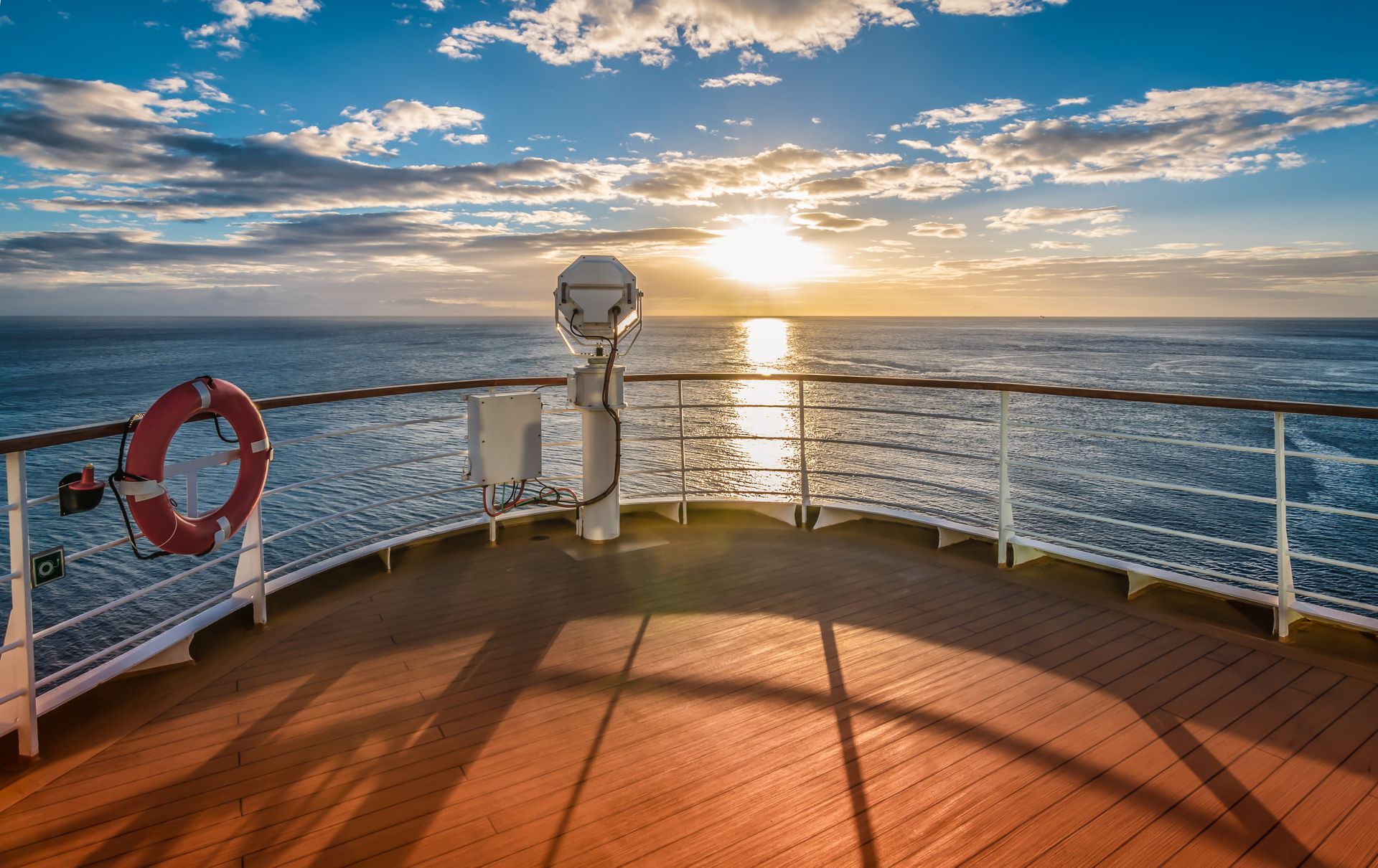 The deck of a cruise ship at sunset, overlooking the calm blue ocean with a life ring mounted on the railing.