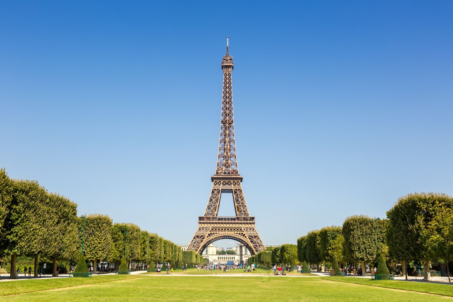 The Eiffel Tower stands in the distance, framed by manicured green trees and a lawn under a clear blue sky in Paris.