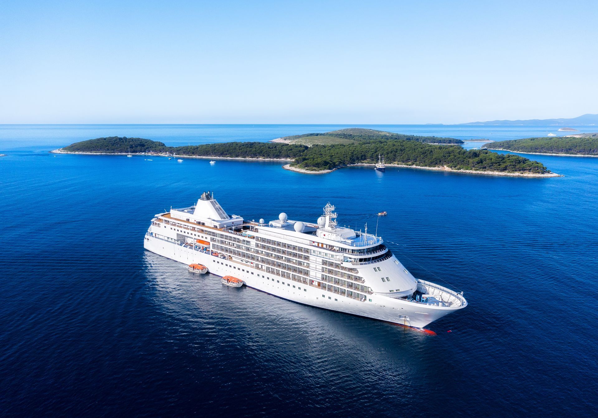 A white cruise ship sailing through calm blue waters near lush green islands on a clear, sunny day.