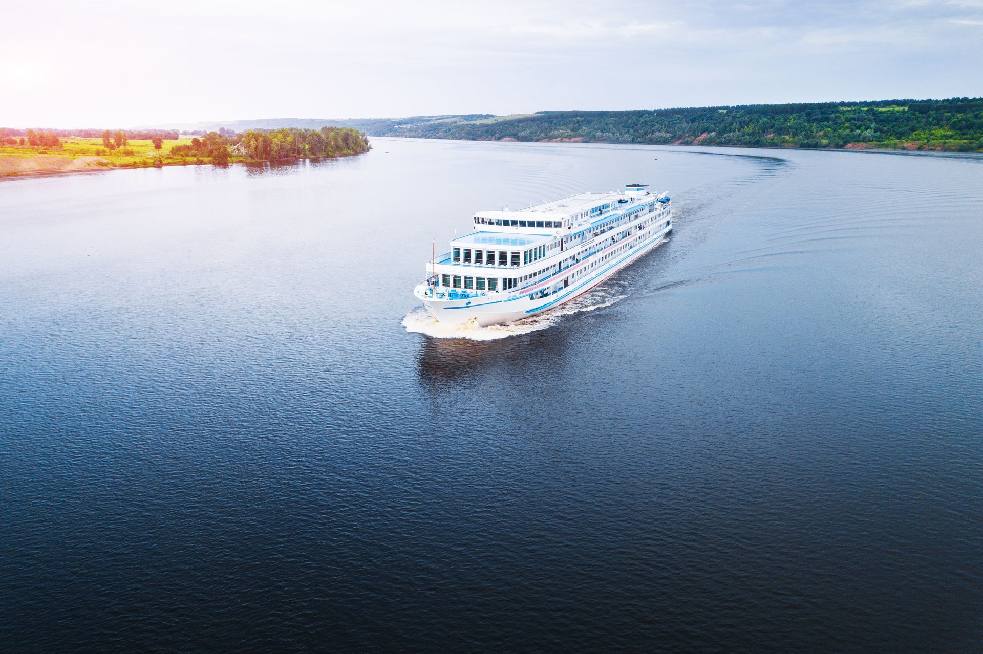 A white cruise ship sails on a wide river with forested banks under a bright, sunny sky. A white cruise ship sails on a wide river with forested banks under a bright, sunny sky.