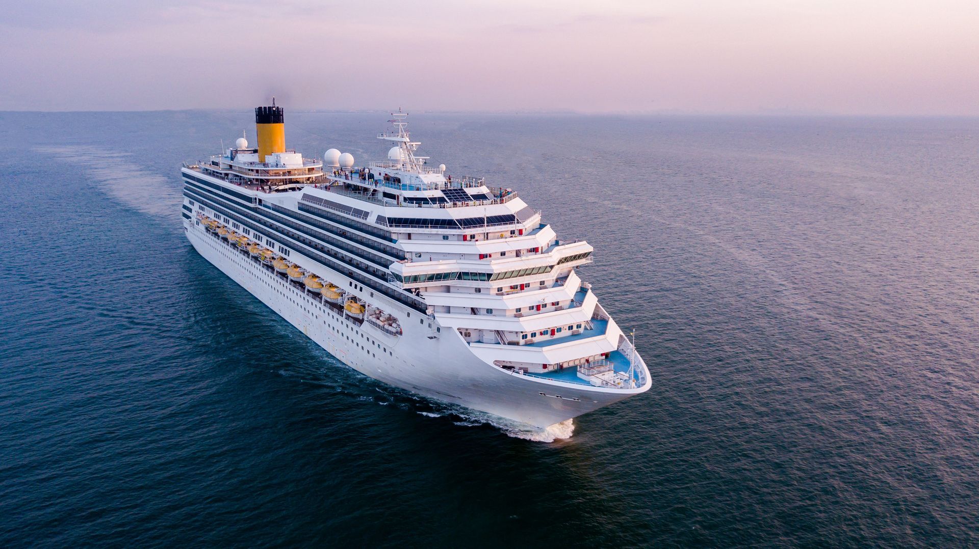 A white cruise ship with a yellow funnel sailing across the blue ocean during a hazy sunset.