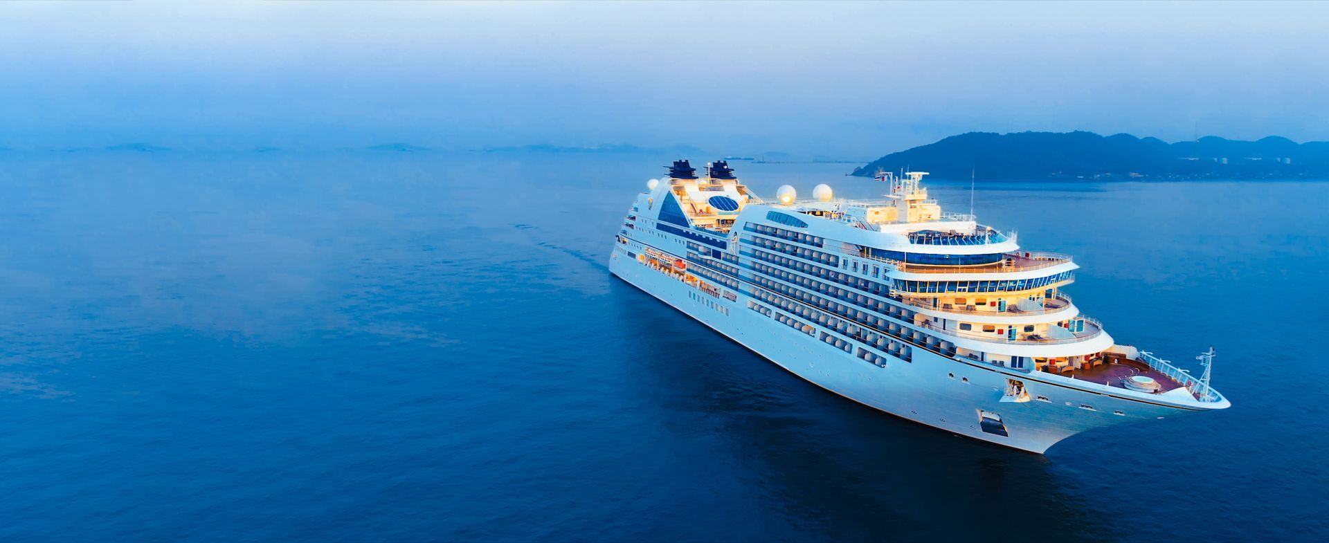 A large white cruise ship sails across a calm, deep blue sea under a soft twilight sky.